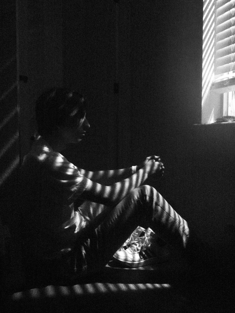 Black and white photo of a thoughtful man sitting by a window with blinds casting shadows.