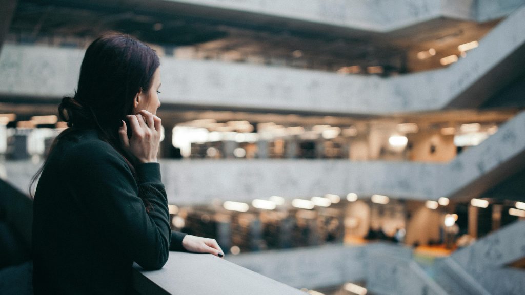 A woman gazes thoughtfully from a balcony in a modern architectural interior space.